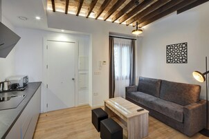 Un espacio habitable moderno con kitchenette, un sofá gris, una mesa de centro de madera y un techo con vigas a la vista.