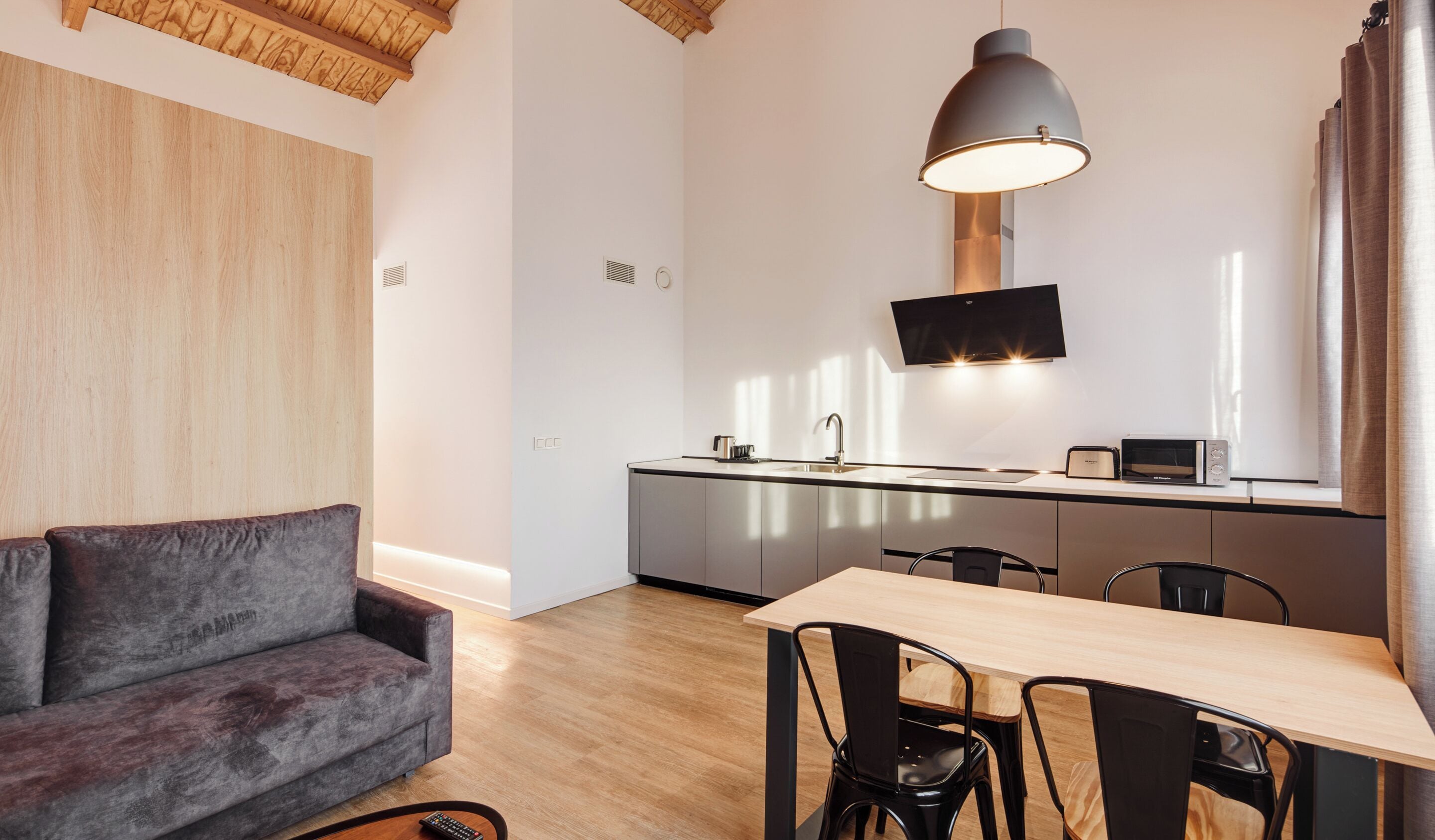 A modern kitchen with a central island, a dining table with chairs, a grey sofa, and a pendant light.