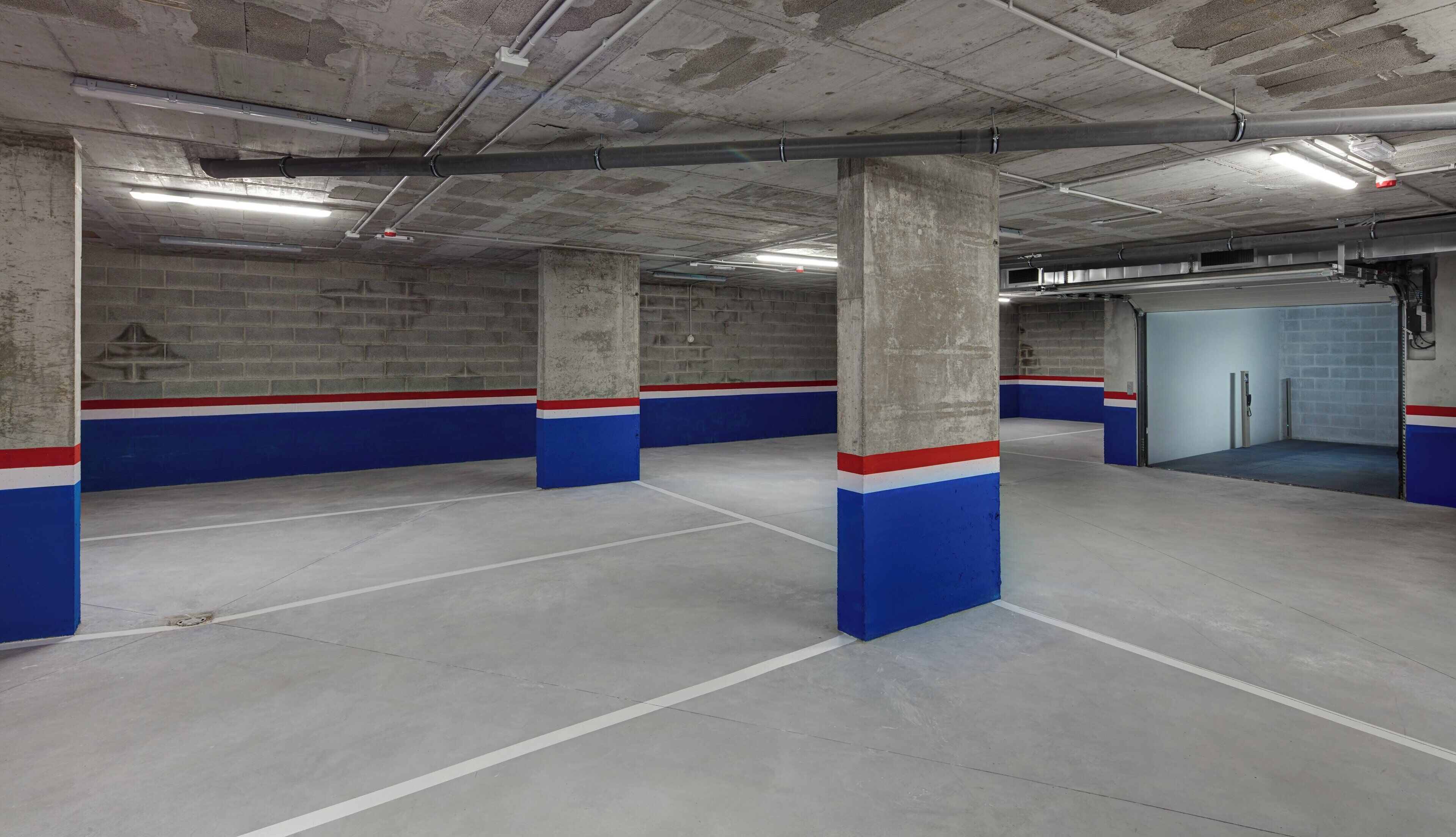 Un aparcamiento con pilares de hormigón, paredes pintadas de azul y rojo, e iluminación fluorescente.