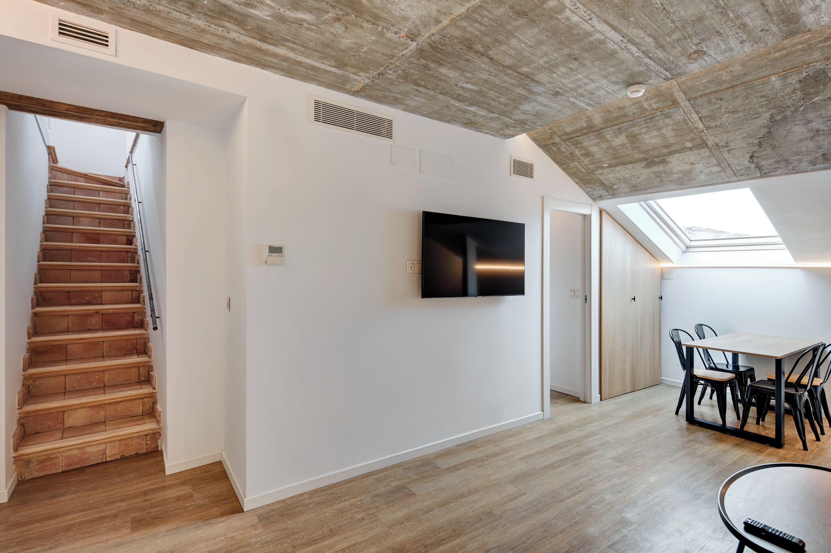 Un interior moderno con una escalera de madera, un televisor de pantalla plana montado en la pared y un tragaluz sobre una zona de comedor.