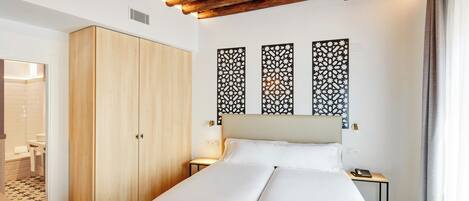 A hotel room with a wooden wardrobe, a bed with white bedding, decorative wall panels, and a bedside table with a lamp.
