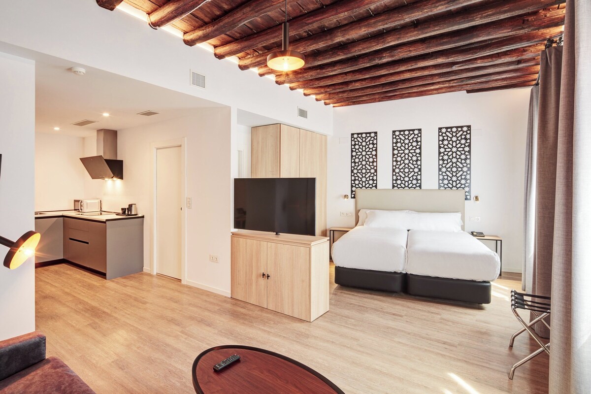 Ein modernes Hotelzimmer mit Holzboden, einem Flachbildfernseher, einem Bett mit weißer Bettwäsche, einem Schreibtisch mit Computer und einem Essbereich.