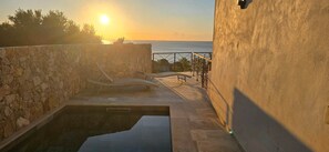 Un mur de pierre, une piscine et une vue sur la mer au coucher du soleil.