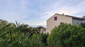 A two-story building with a beige facade and purple shutters, surrounded by lush greenery and a clear sky.