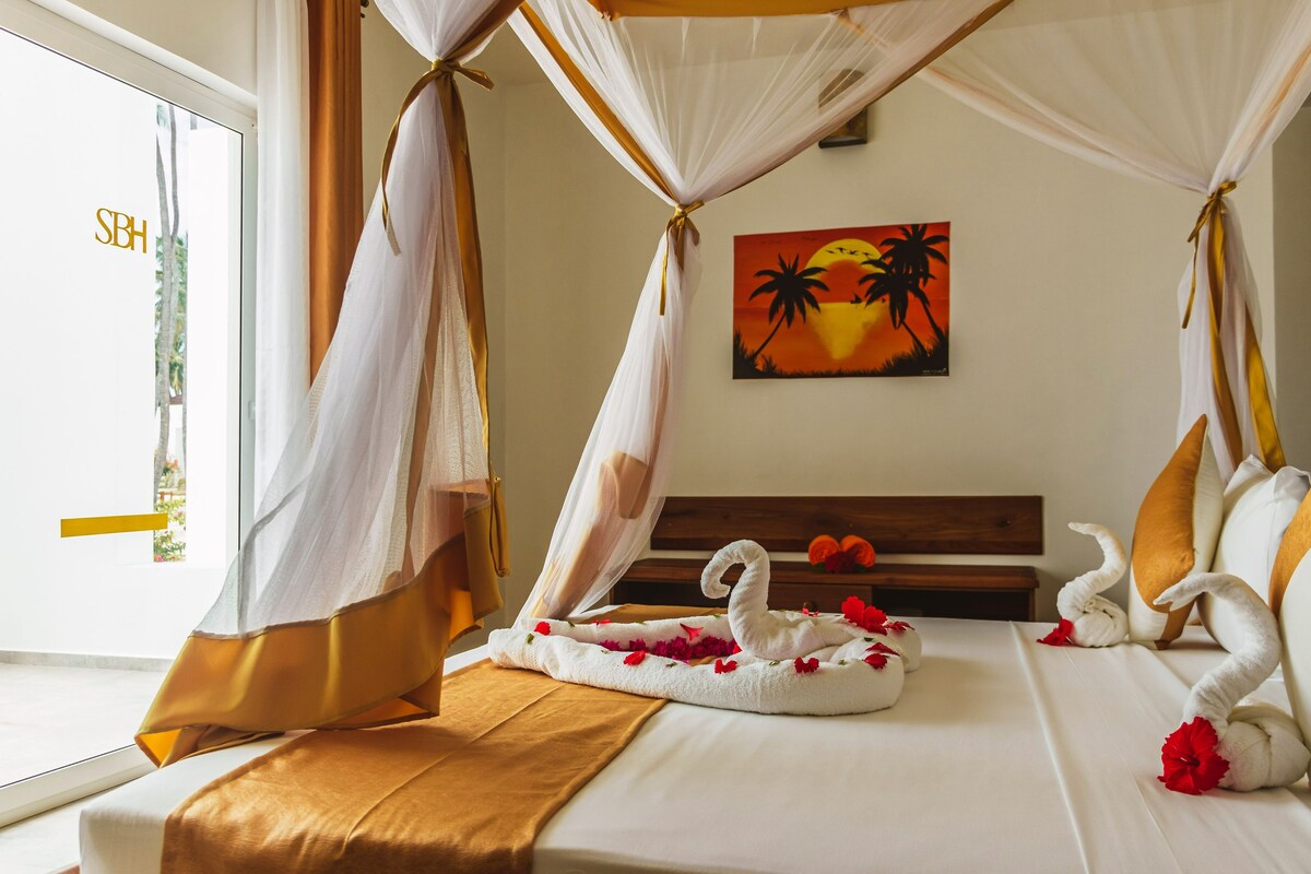A hotel room with a bed, a painting of palm trees and a sunset, and a swan towel decoration.