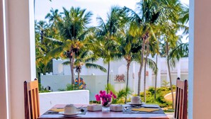 A dining table with chairs, a vase with pink flowers, and a view of palm trees and a white building. - SBH Kilindini Resort (Matemwe)