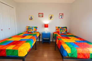 Habitación con dos camas, cada una con una colcha de colores con motivos de LEGO, una mesita de noche azul pequeña con una lámpara roja y una puerta blanca.