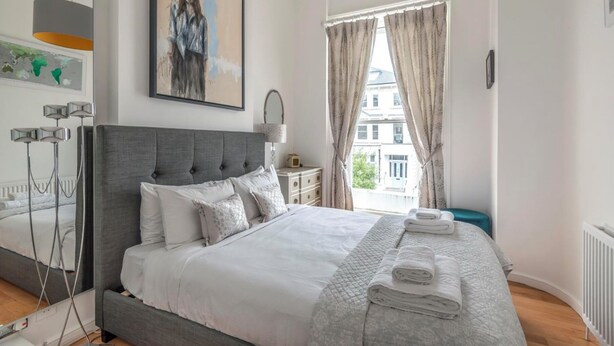 Une chambre à coucher bien rangée, avec un grand lit, une table de chevet, une lampe et une vue sur l’extérieur.