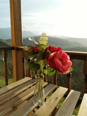 A wooden table with a clear glass vase containing red flowers and green foliage, set against a backdrop of a mountainous landscape. - Valle la Fuente (Cabrales)