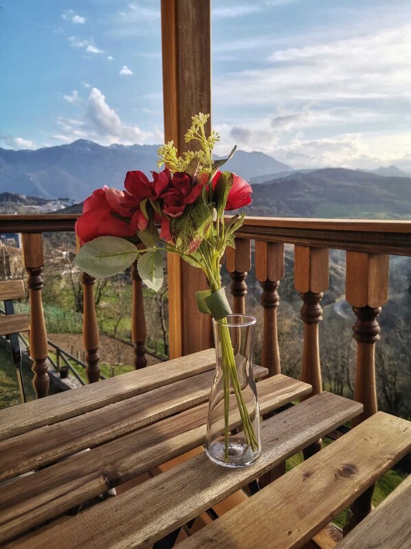 A wooden table with a clear glass vase containing red flowers and green leaves, set on a balcony with a mountain view. - Valle la Fuente (Cabrales)