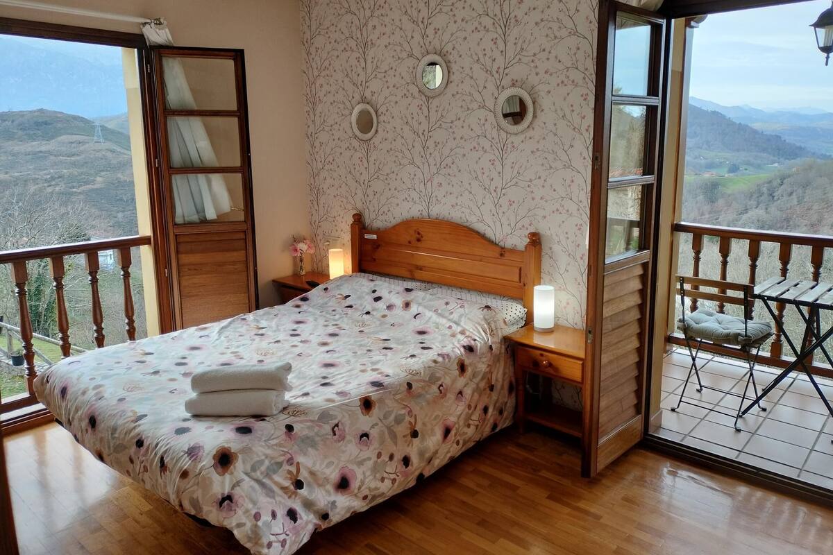 A bedroom with a wooden bed, a nightstand, a bedside lamp, and a view of the mountains through a balcony.