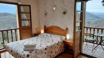 A bedroom with a wooden bed, a nightstand, a bedside lamp, and a view of the mountains through a balcony.