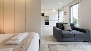 Un dormitorio moderno con una cama, un sofá gris y vistas a una cocina y un balcón.