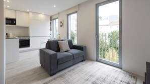 Un apartamento moderno con un sofá gris, una cocina con armarios blancos y un amplio ventanal corredera a un balcón.