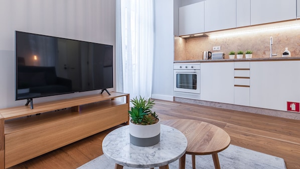 Un salon moderne avec un téléviseur à écran plat posé sur un meuble en bois, une table en marbre agrémentée d’une plante en pot, et une cuisine avec des meubles blancs et un four encastré.