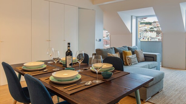 Een eetruimte met een tafel gedekt voor vier personen, inclusief borden, glazen en bestek. Op tafel staat een fles wijn. De kamer heeft uitzicht op een stadsbeeld door een raam.