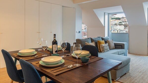 Un coin repas avec une table dressée pour quatre personnes, comprenant des assiettes, des verres et des couverts. Une bouteille de vin est posée sur la table. La pièce offre une vue sur le paysage urbain grâce à une fenêtre.