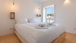 Een moderne slaapkamer met een groot bed, witte lakens, een klein nachtkastje en een raam met uitzicht op gebouwen.