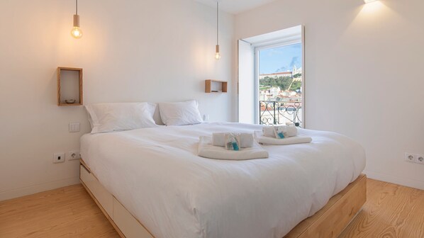 Une chambre moderne avec un grand lit, du linge de lit blanc, une petite table de chevet et une fenêtre donnant sur des immeubles.