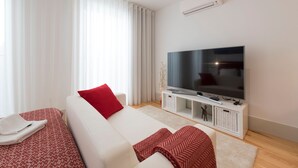 Een moderne woonkamer met een witte bank, rode kussens, een flatscreen televisie op een witte dressoir, en een groot raam met dunne gordijnen.