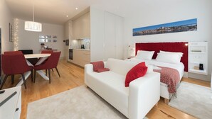 Een moderne hotelkamer met een witte bank, rode kussens, een bed met witte beddengoed, een rood hoofdeinde, een zithoek met een rode stoel, een keukenhoek en een stadszicht op de muur.
