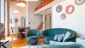 Un soggiorno moderno con un divano color petrolio, una zona pranzo con tavolo in legno e una scala che conduce a un piano superiore.