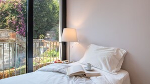 Ett hotellrum med en säng, ett nattduksbord med en bok och en kopp, en lampa och utsikt över grönområden samt en balkong.