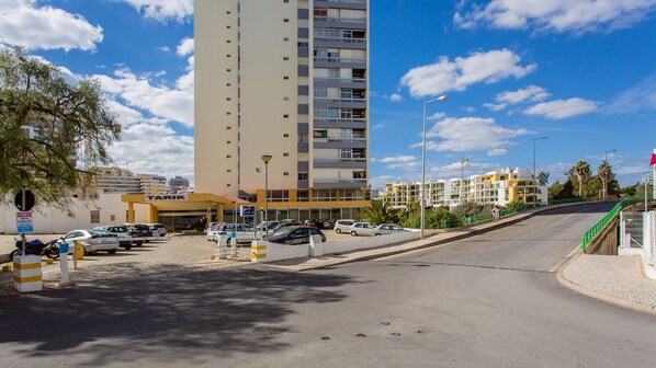 Ein Parkplatz mit mehreren Autos, ein hohes Gebäude mit Balkonen und ein klarer, blauer Himmel.