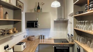 Una cocina moderna con horno empotrado, microondas y un lavabo. Hay estantes con cristalería y una lámpara colgante.