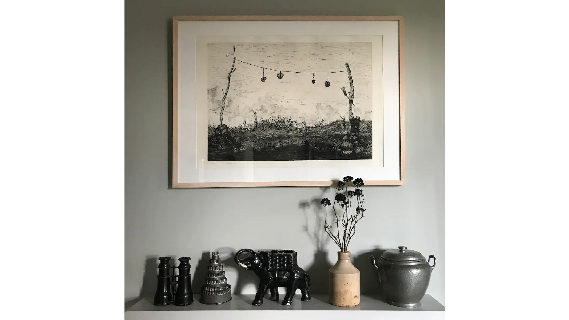 Ett inramat svartvitt fotografi som föreställer ett landskap med en ljusslinga, en liten byggnad och ett träd. Under bilden finns svarta dekorativa föremål, bland annat en vas med grenar, en keramikelefant och en metallkruka.