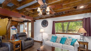 A spacious living room with a high ceiling, a large window with a view of trees, a leather sofa, a coffee table, and a fireplace. - Enchanting Lake Texoma Cabin with Hot Tub (Pottsboro)