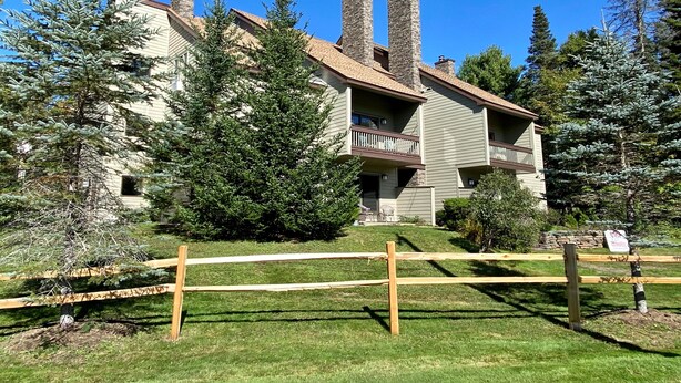 Un edificio de dos pisos con un balcón en el segundo nivel, rodeado de árboles y una valla de madera.