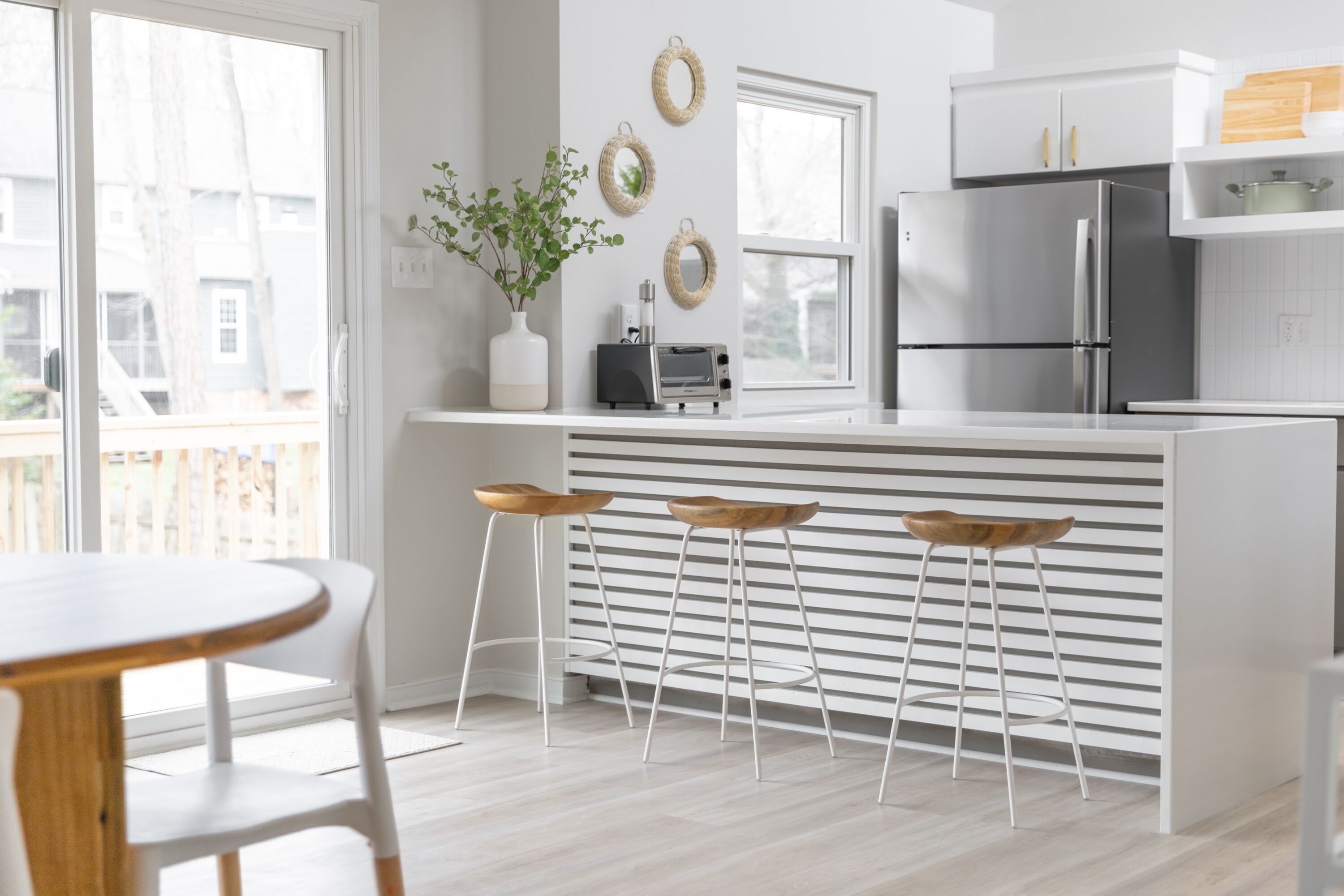 Une cuisine moderne avec des meubles blancs, un réfrigérateur en acier inoxydable, un four encastré et un petit îlot central avec des tabourets.