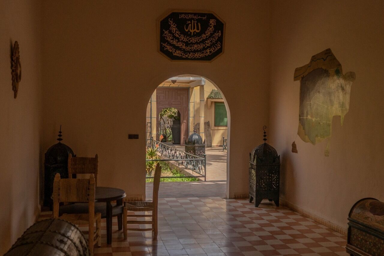 Un corridoio interno con pavimento a scacchi, un tavolo in legno con sedie, una decorazione a parete e una vista attraverso un arco su un'area esterna con una fontana e vegetazione.