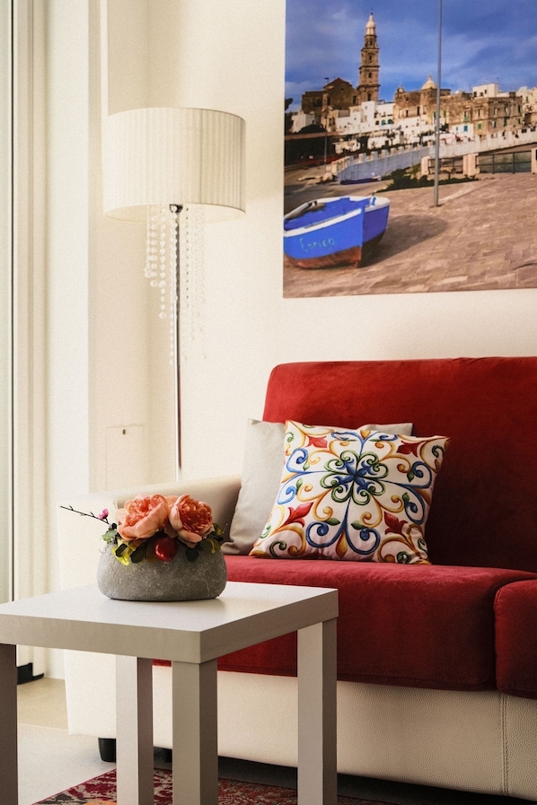 Un salon avec un canapé rouge, une table blanche, un vase décoratif avec des fleurs, une lampe et un tableau accroché au mur représentant une ville côtière.