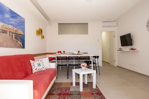 Un salon moderne avec un canapé rouge, une table à manger avec des chaises, une petite table blanche ornée d’un vase et un tableau représentant un paysage côtier.