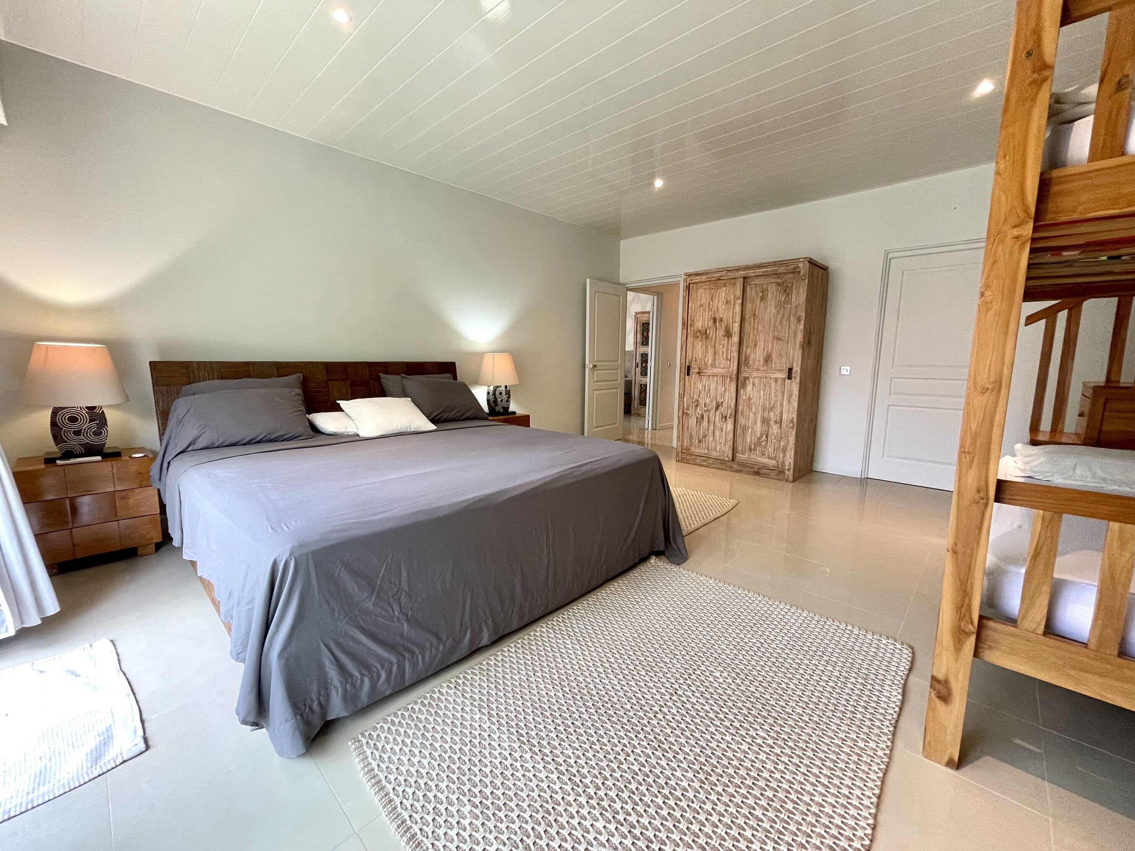 Une chambre avec un grand lit, une tête de lit en bois, deux tables de chevet avec des lampes, une armoire et un lit superposé dans un coin.