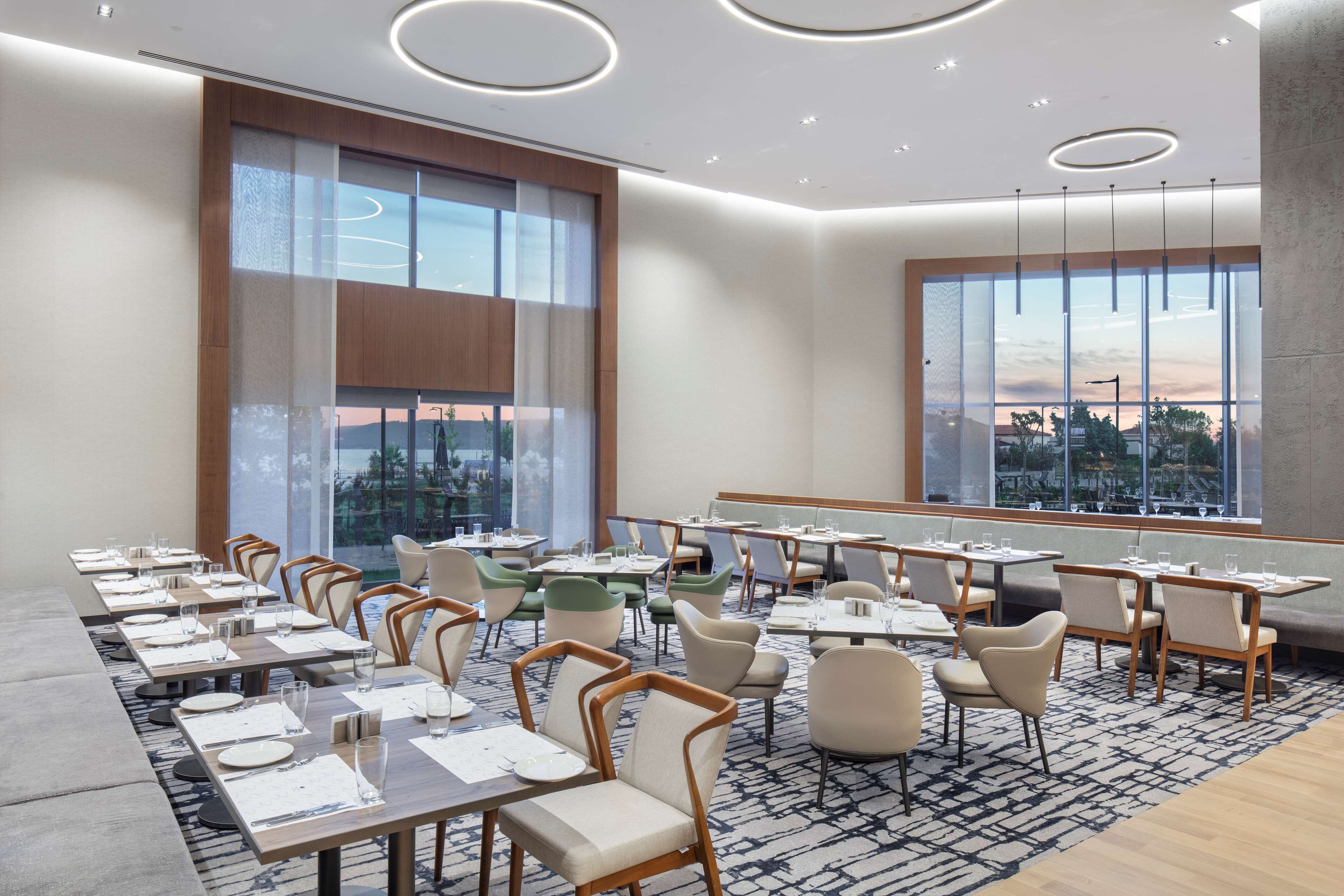 A modern dining area with long tables, chairs, and large windows offering a view of the cityscape.