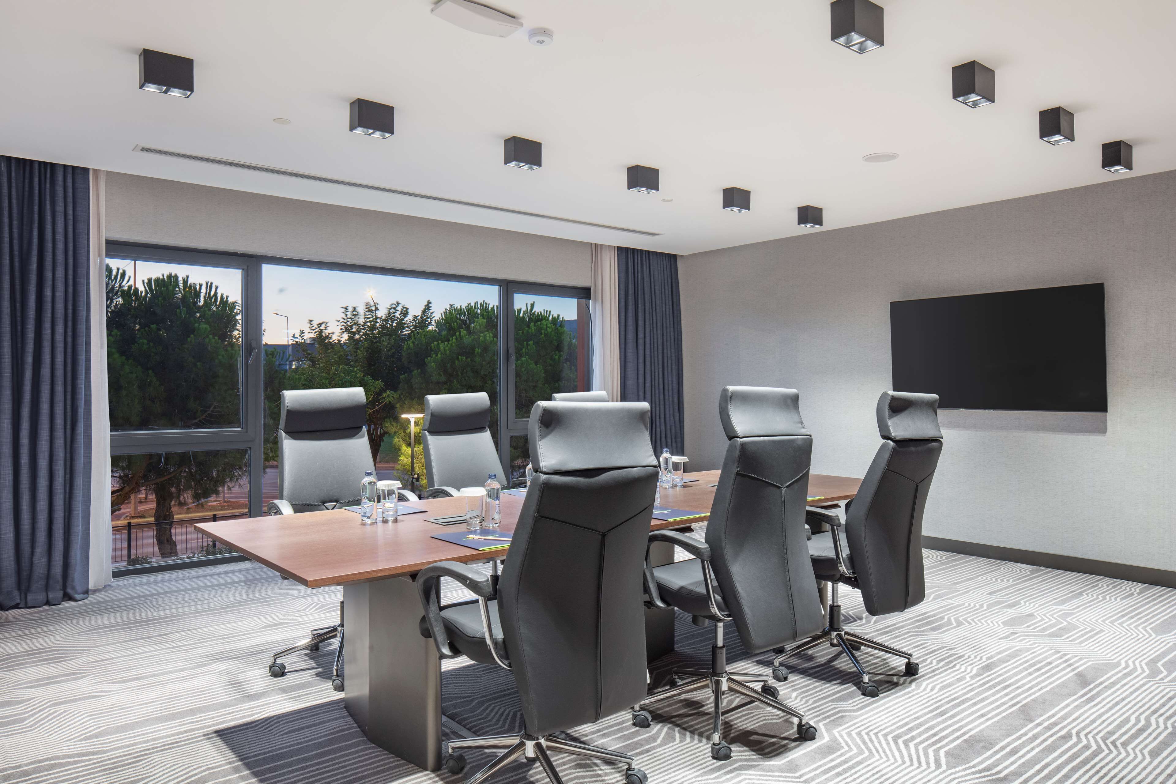Un salón de conferencias con una gran mesa, cuatro sillas, una pantalla de proyector y vistas a árboles a través de amplias ventanas.