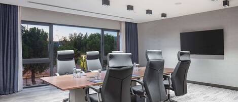 Un salón de conferencias con una gran mesa, cuatro sillas, una pantalla de proyector y vistas a árboles a través de amplias ventanas.