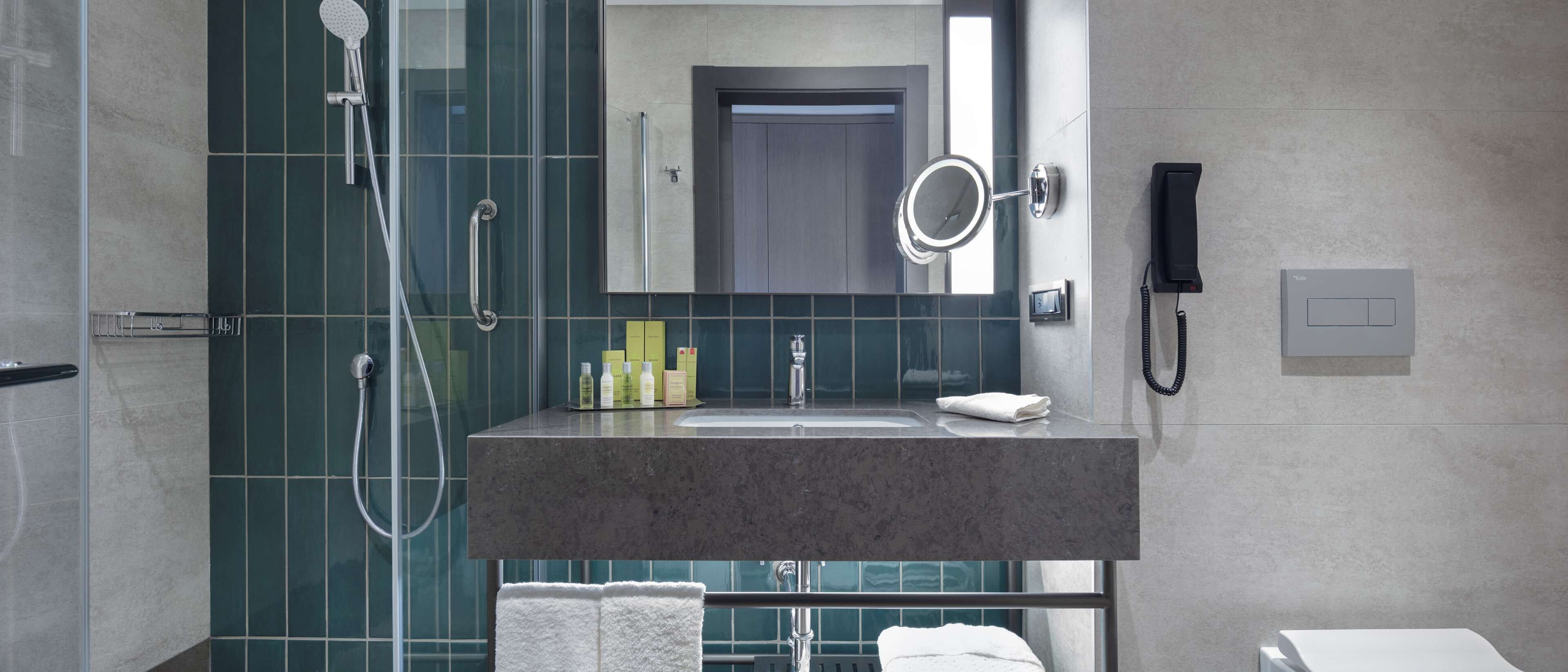 A modern bathroom with a shower, a sink with toiletries, a mirror, and a phone.