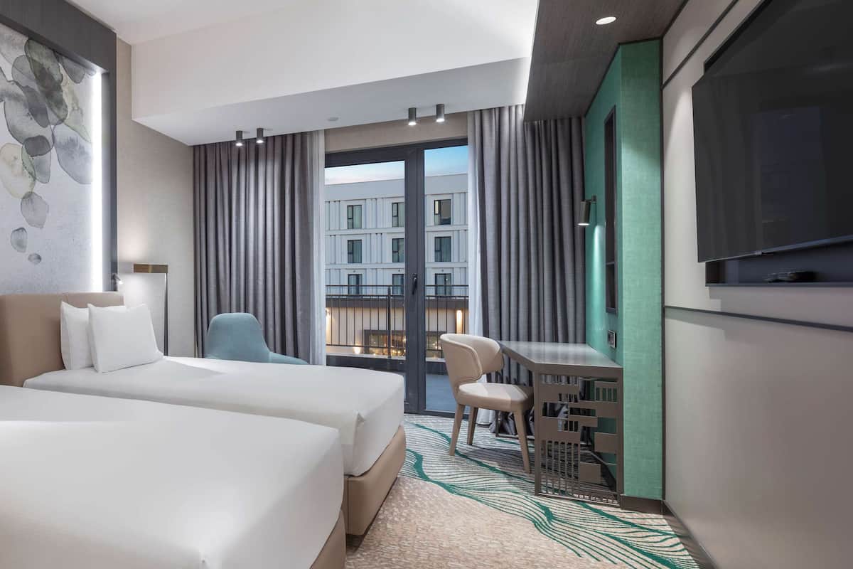 A hotel room with two beds, a desk, a chair, a TV, and a view of the cityscape.
