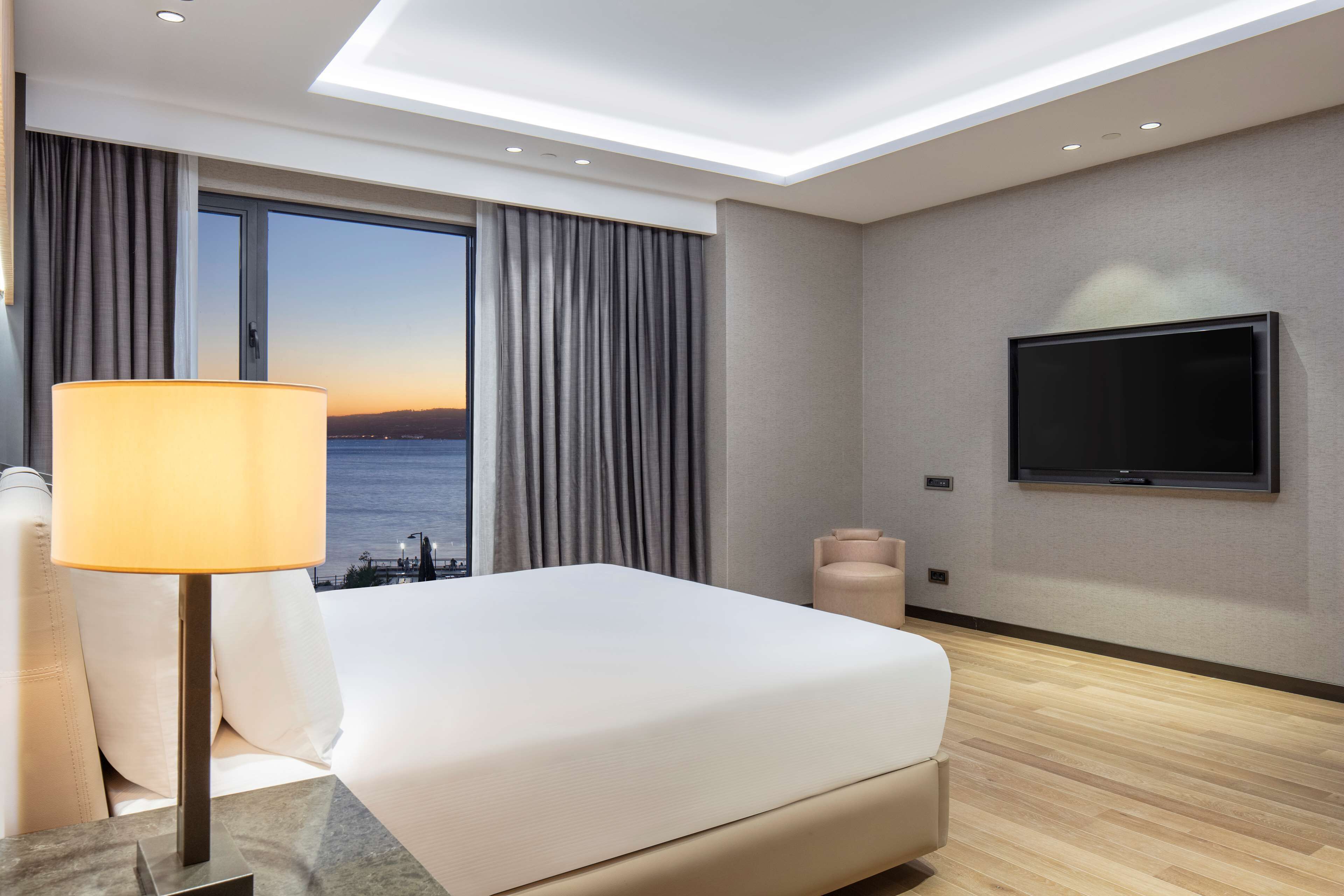 A modern hotel room with a large bed, a flat-screen TV, a lamp, and a view of the sea.