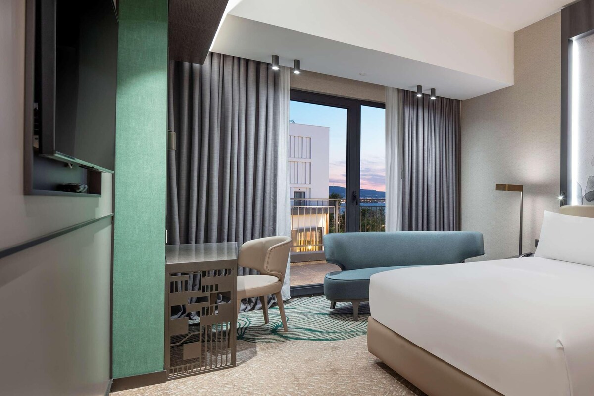 Habitación de hotel con una cama grande, un escritorio con silla, un sofá azul y vistas a la ciudad a través de la ventana.