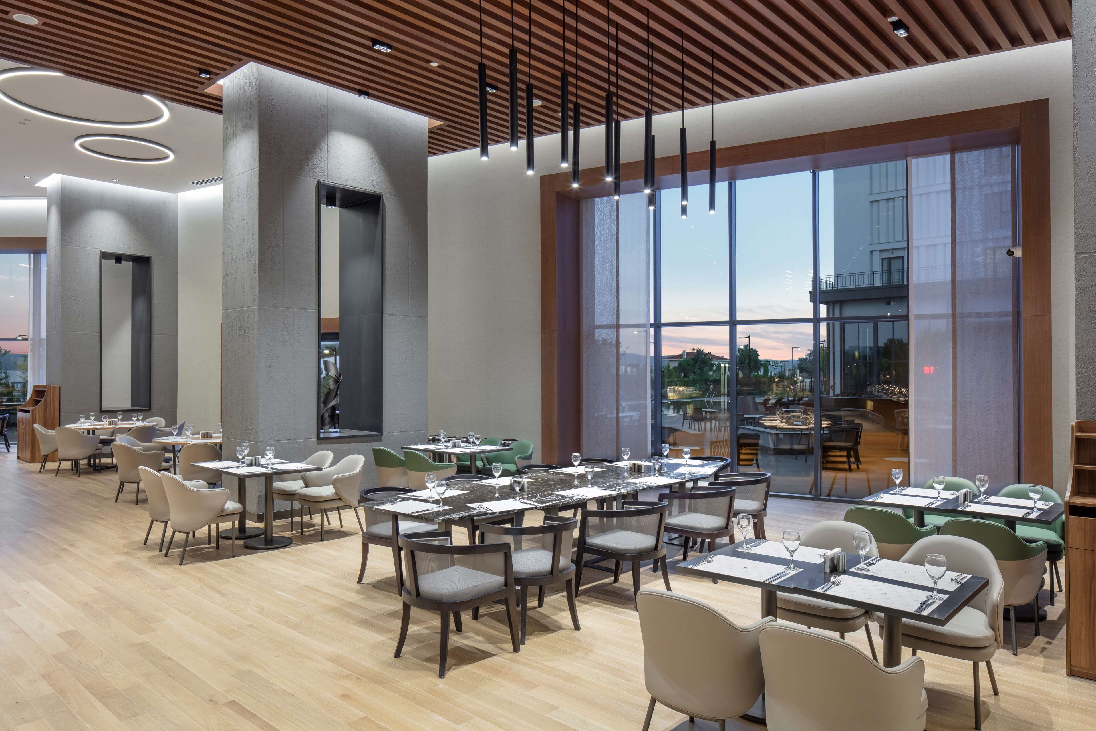 A modern restaurant with multiple dining tables and chairs, large windows offering a view of the cityscape, and a wooden ceiling with hanging lights.