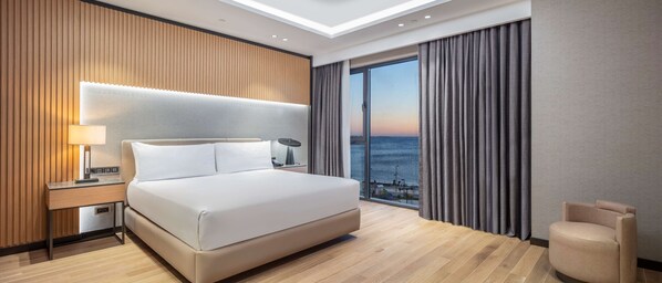 A modern hotel room with a large bed, a nightstand with a lamp, a view of the sea through the window, and a wooden wall paneling. - Doubletree By Hilton Canakkale (Çanakkale)