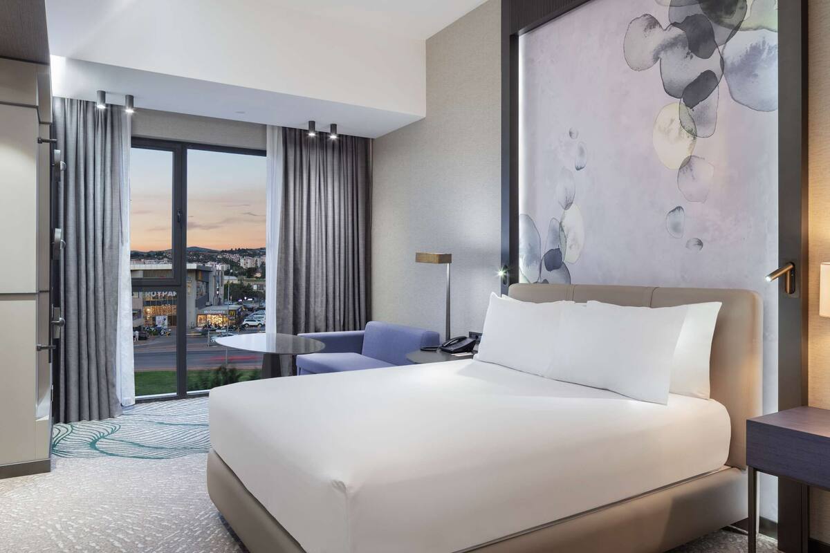 A modern hotel room with a large bed, a desk, a chair, and a view of the city through a large window.