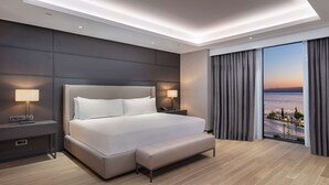 Una habitación de hotel moderna con una cama grande, mesitas de noche con lámparas, un banco y vista al mar a través de ventanales de gran altura.
