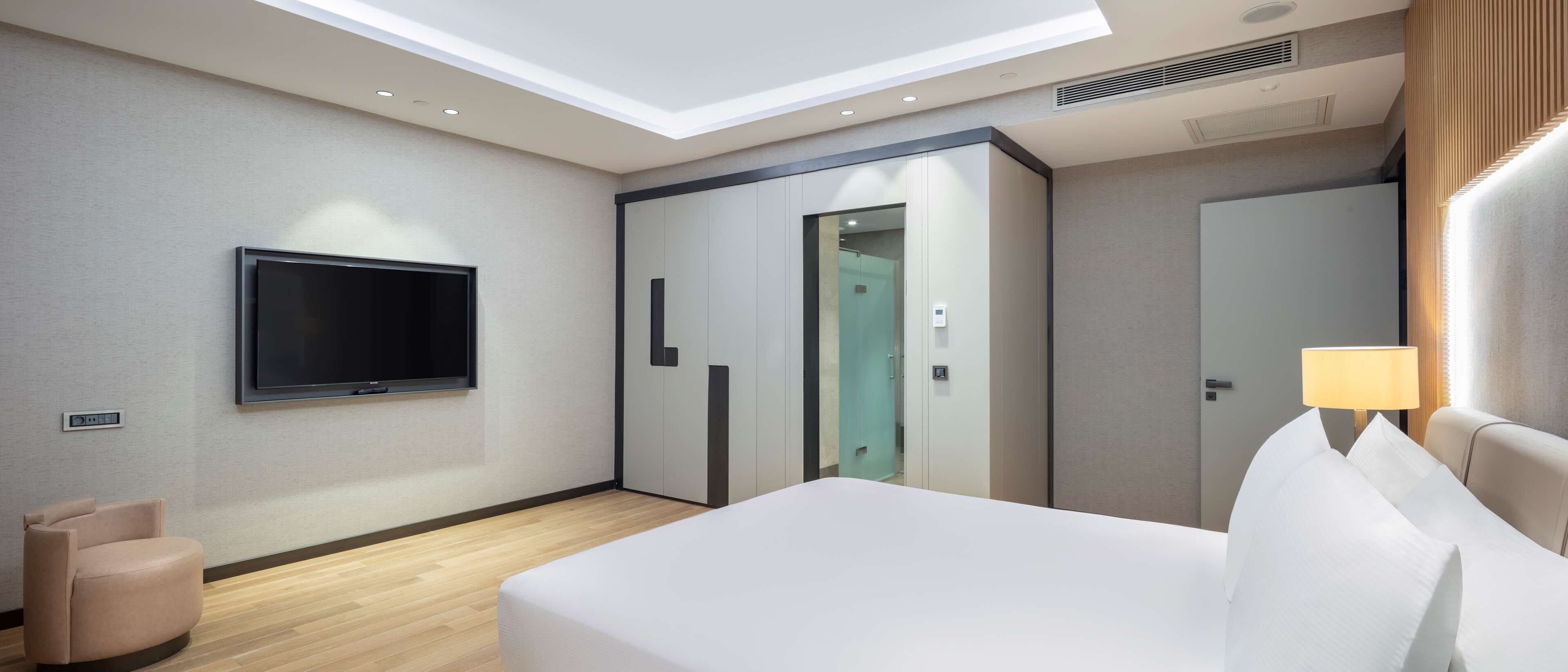 Una habitación de hotel moderna con una televisión de pantalla plana montada en la pared, una cama con ropa de cama blanca, una lámpara de noche y un armario con puertas correderas.