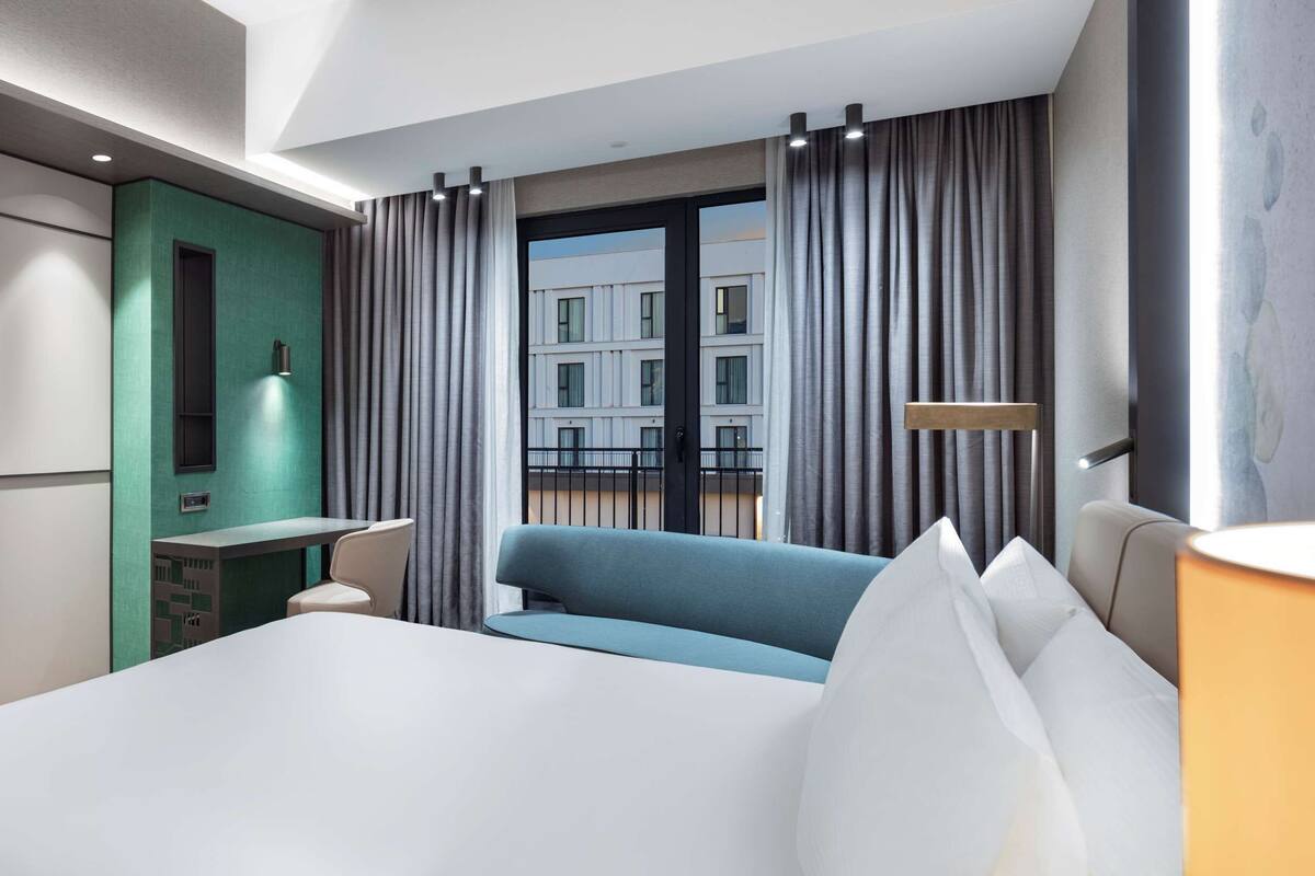 A modern hotel room with a large bed, a television mounted on the wall, a small table, and a view of the city through the window.